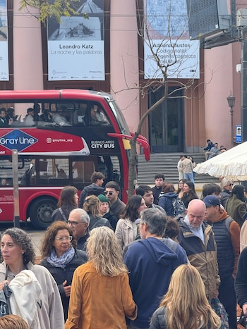 Personas caminando en la calle frente a un autobús turístico rojo y un edificio de pared rosada con carteles de arte