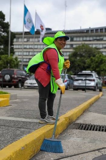 Mujer con chaleco reflectante verde, gorra y guantes barre con una escoba azul junto a un bordillo amarillo y vehículos estacionados