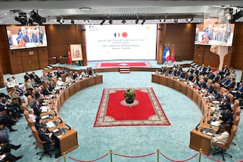 Salón de conferencias con mesas de madera semicirculares y delegados sentados, pantalla central con texto, dos pantallas laterales, banderas de Marruecos y Francia, alfombras rojas
