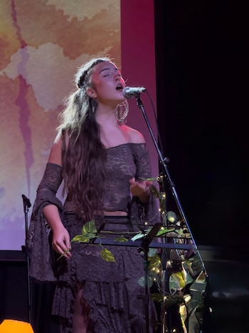Una persona con cabello largo toca una guitarra acústica con los ojos cerrados frente a un micrófono en un escenario iluminado