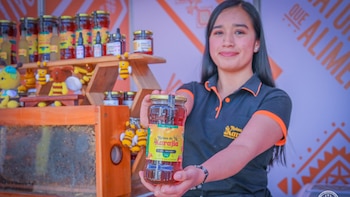 Mujer sostiene un frasco grande de miel con la etiqueta "Reina de Karajía" en un puesto con productos apícolas, abejas vivas y juguetes de abeja