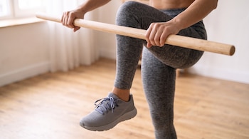 Persona con mallas y zapatillas deportivas que sostiene una barra de madera horizontal con ambas manos y levanta una pierna flexionada.
