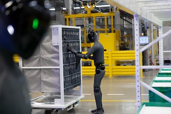 Robot humanoide oscuro manipulando un estante blanco con componentes en una fábrica. Otro robot borroso en primer plano. Maquinaria amarilla y contenedores verdes