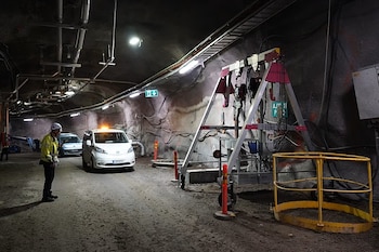 Trabajador con casco y chaleco amarillo junto a una furgoneta blanca en un túnel subterráneo industrial. Se ven tuberías, cables y equipos de seguridad