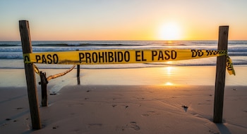 Cinta amarilla con la frase Prohibido el paso extendida entre dos postes de madera en una playa con arena, mar, olas y un atardecer
