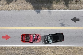 Vista aérea de dos automóviles colisionados, uno rojo y uno negro, en una carretera de doble carril con línea amarilla. Flechas direccionales en el asfalto. Vegetación seca al borde
