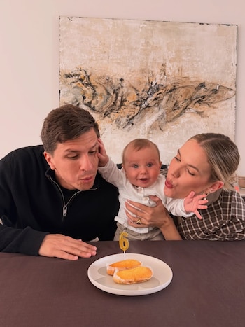 Eva Bargiela y Gianluca Simeone con su hijo Faustino, celebrando sus seis meses con una vela "6" y dos pasteles en una mesa oscura, sonriendo