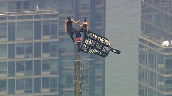 Dos personas vestidas de negro con máscaras en la cima de una torre metálica, sosteniendo una pancarta negra con letras blancas sobre un edificio y agua