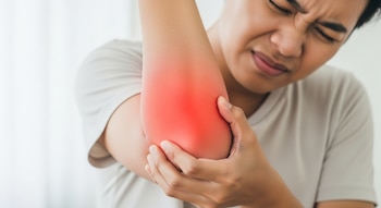 Persona con expresión de dolor sostiene su codo derecho, que muestra una zona rojiza brillante indicando molestia. Viste camiseta clara sobre fondo blanco.