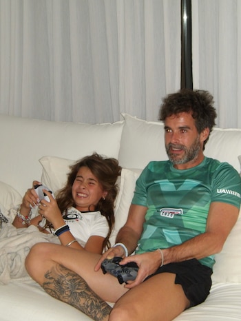 Un hombre y una niña están sentados en un sofá blanco con dos controles de videojuegos. La niña sonríe y el hombre lleva una camiseta verde