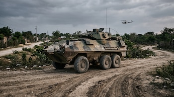 Vehículo blindado militar de seis ruedas sobre un camino de tierra con huellas marcadas, vegetación baja y un helicóptero en el cielo.