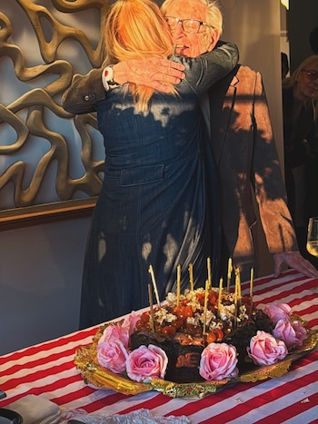 Una mujer rubia abraza a un hombre con gafas. Una torta de chocolate con velas, cerezas y rosas rosas sobre un mantel a rayas