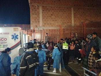 Vista nocturna de personal médico y de policía trasladando un paciente en camilla hacia una ambulancia en Cusco, con un grupo de personas observando detrás de una valla metálica