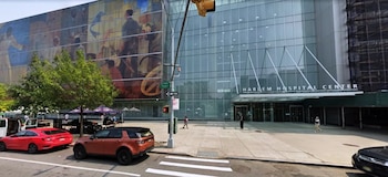 Fachada de vidrio del Harlem Hospital Center con mural, entrada principal, vehículos estacionados en la calle, personas caminando, semáforo y andamios