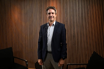Hombre de pie con blazer oscuro, camisa clara, pantalón gris, micrófono de solapa, frente a una pared de listones de madera, con dos sillas oscuras a los lados