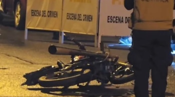 Una motocicleta oscura tirada en el asfalto de noche, cerca de barreras amarillas con "ESCENA DEL CRIMEN" y un oficial de policía de espaldas