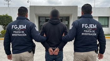 Un hombre esposado, visto de espaldas con el rostro borroso, es flanqueado por dos agentes de la FGJEM de espaldas frente a un edificio.