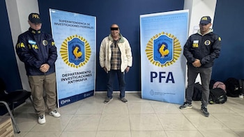 Un hombre de mediana edad con el rostro censurado flanqueado por dos agentes de la PFA uniformados. Detrás, dos estandartes de la Policía Federal Argentina