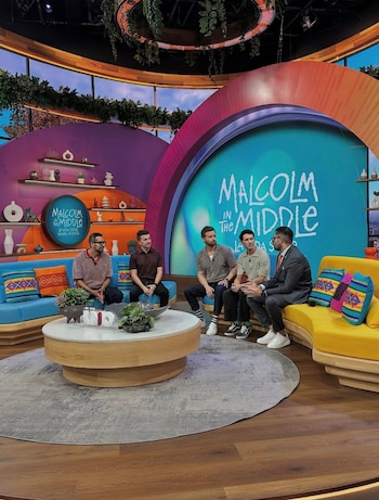 Cinco hombres sentados en un colorido set de televisión. Dos sofás, uno azul y otro amarillo, flanquean una mesa de centro redonda. El fondo muestra el logo 'Malcolm in the Middle'
