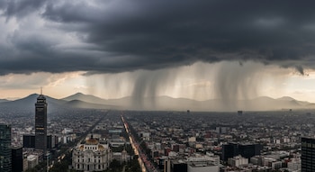 Panorámica de una ciudad con rascacielos y edificios bajos, una avenida con tráfico vehicular, montañas al fondo y nubes de tormenta con lluvia visible.