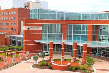 Edificio de ladrillo rojo y vidrio con letrero "Suburban Hospital", entrada principal, bandera estadounidense, vehículos y paisajismo en el exterior