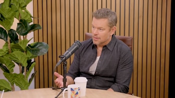 Matt Damon sentado a una mesa de madera con un micrófono negro, dos tazas, una planta verde y una pared de listones de madera clara de fondo