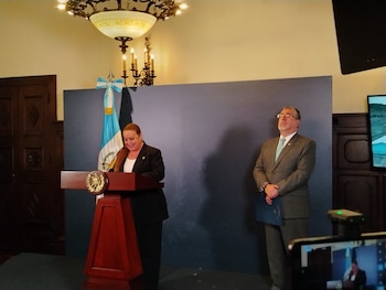 Mujer en traje oscuro habla desde un podio con el escudo de Guatemala, frente a una bandera azul y blanca. Un hombre de traje claro está a su derecha