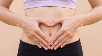 Torso y abdomen de una persona vestida con top lavanda que ubica sus manos en forma de corazón a la altura del ombligo.