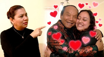 Una mujer a la izquierda señala con el dedo mientras mira a una pareja que se abraza felizmente, rodeada de corazones flotantes rojos y rosas.