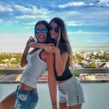 Dos mujeres, una sonriendo ampliamente con gafas de sol azules y otra con gafas oscuras, se abrazan en un balcón con vista a una ciudad y un cielo nublado