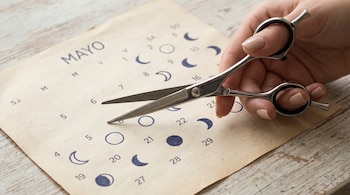 Primer plano de una mano sosteniendo tijeras de peluquería sobre un calendario de papel beige del mes de Mayo con símbolos de fases lunares y números.