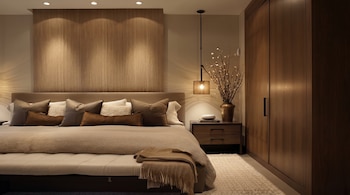 Vista frontal de un dormitorio moderno en tonos neutros con una cama grande, almohadas, banco, mesita de noche, lámpara colgante y armario de madera oscura.