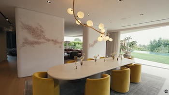 Comedor con mesa ovalada, varias sillas amarillas, candelabro ramificado, paredes con decoración floral y ventanales con vista a un jardín y piscina