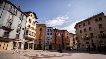 Una plaza empedrada rodeada de edificios de colores con balcones y locales comerciales, por la que caminan algunos peatones.