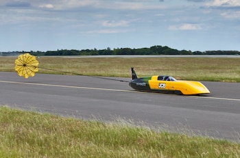 Coche aerodinámico amarillo y negro con paracaídas amarillo brillante desplegado en una pista, con hierba y árboles al fondo bajo cielo nublado