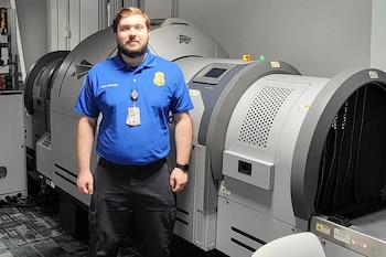 Hombre con barba y uniforme azul, con credencial de oficial, de pie junto a una máquina grande de inspección de equipaje con banda transportadora