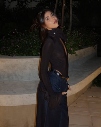 Cami Mayan sonríe sosteniendo un ramo de flores envuelto en papel blanco, vestida con top oscuro, jeans y cinturón dorado, frente a unas escaleras de madera