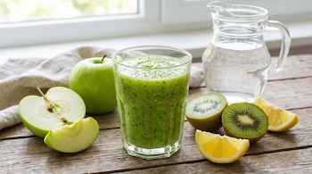 Un vaso de batido verde, manzanas verdes (una entera, una partida), kiwis partidos, rodajas de limón, jarra de agua, paño y mesa de madera.