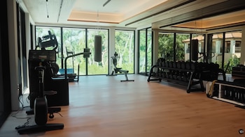 Interior de un gimnasio con grandes ventanales, suelo de madera clara, bicicleta elíptica, saco de boxeo, máquina de remo y estante con mancuernas