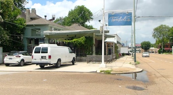 Una van Ford E-Series blanca estacionada junto a un SUV blanco. Un edificio con el letrero 'Tamboli's Pasta & Pizza' y asientos. Calle con charco y vehículos al fondo