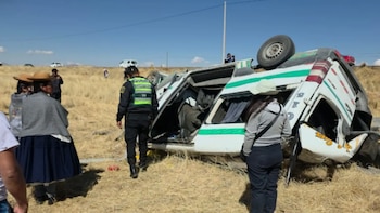 Una minivan blanca con franjas verdes volcada en un campo seco junto a un agente de policía y varias personas que observan el vehículo