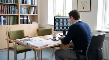 Profesional médico de espaldas revisando imágenes de resonancia magnética cerebral en un monitor. El escritorio tiene documentos, bolígrafos y una taza. Fondo con estanterías de libros.