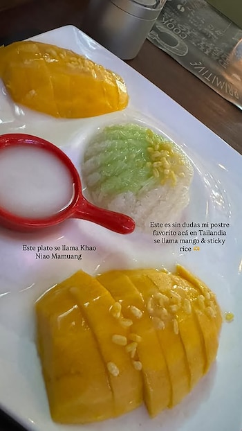 Un plato blanco presenta dos mitades de mango amarillo brillante en rodajas, arroz pegajoso blanco y verde, y un tazón rojo con salsa blanca de coco