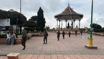 Varios soldados armados caminan por una plaza central pavimentada, con un kiosko de techo rojo y árboles frondosos. Dos civiles están sentados a la izquierda