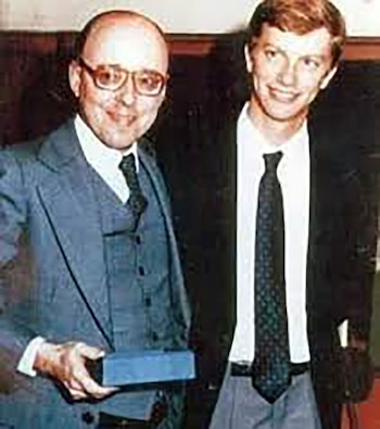 Dos hombres sonríen para la cámara: Jaime Guzmán, calvo y con gafas, en traje gris, sostiene un objeto rectangular azul; a su lado, José Antonio Kast, joven con cabello castaño y traje oscuro