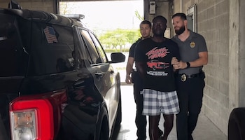 Un hombre de tez oscura con camiseta negra y pantalones a cuadros es escoltado por dos agentes de policía junto a un vehículo policial negro