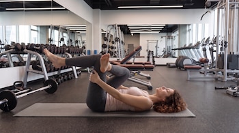 Una mujer acostada en el suelo de un gimnasio sobre una colchoneta realiza un estiramiento de piernas junto a pesas y máquinas.