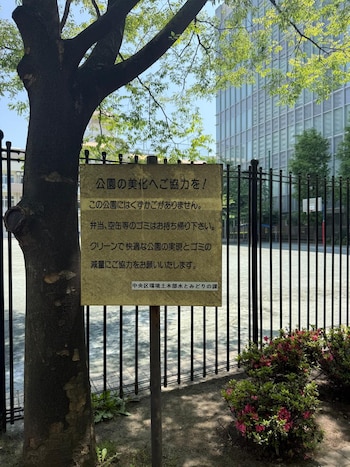 Cartel metálico con texto en japonés fijado a un árbol de tronco oscuro y hojas verdes. Detrás, una valla negra y un edificio moderno. Arbustos con flores rosas en primer plano