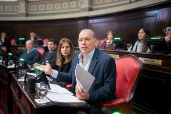 Un hombre vestido con traje habla en un podio con micrófono, mientras sostiene papeles. Otras personas están sentadas en escritorios con micrófonos