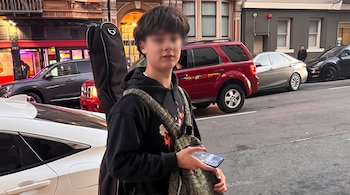 Joven con la zona de los ojos borrosa, una funda negra a la espalda y un teléfono en la mano, de pie en una calle con vehículos.